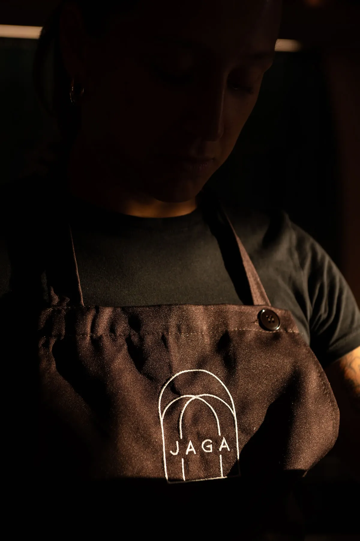 Una cuoca di Jaga nella penombra calda della cucina, il logo ad archi ricamato sul grembiule