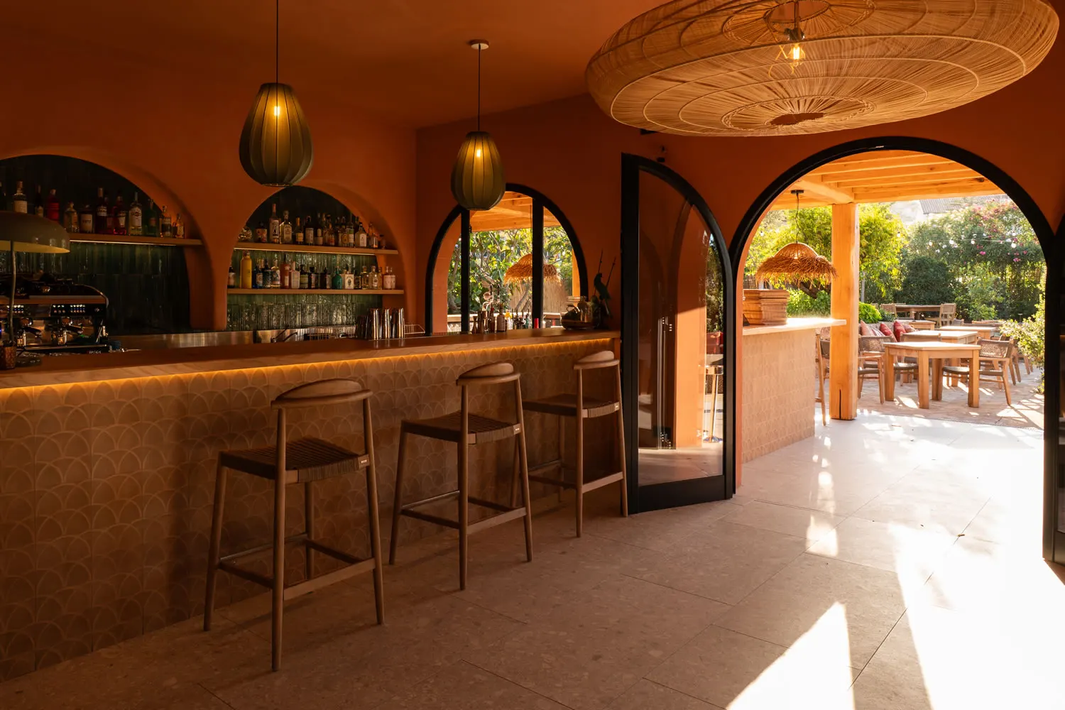 Il bancone interno di Jaga tra nicchie ad arco, lampade in vimini e la terrazza assolata oltre la vetrata